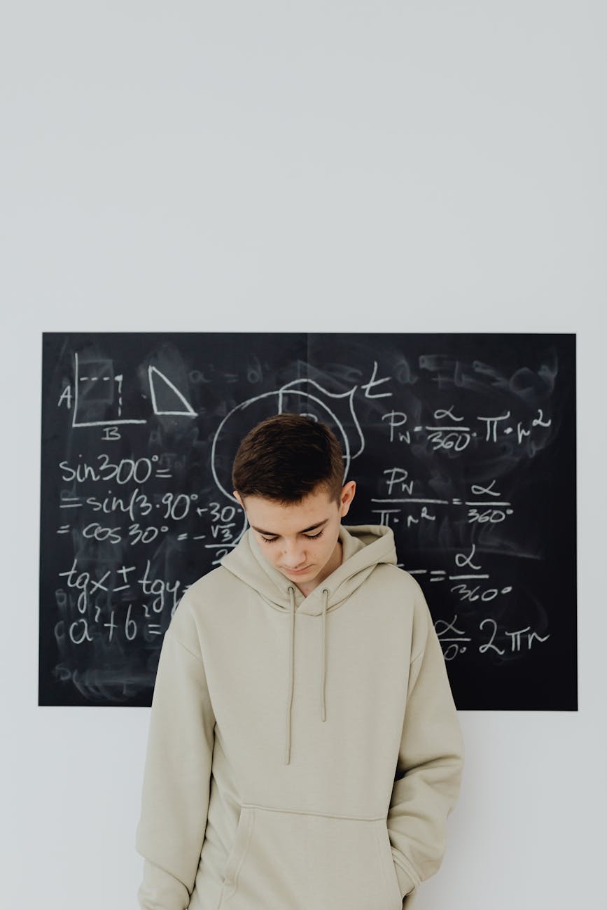 Teen boy in hoodie standing in front of a chalkboard filled with math equations, looking down, reflecting education challenge.
