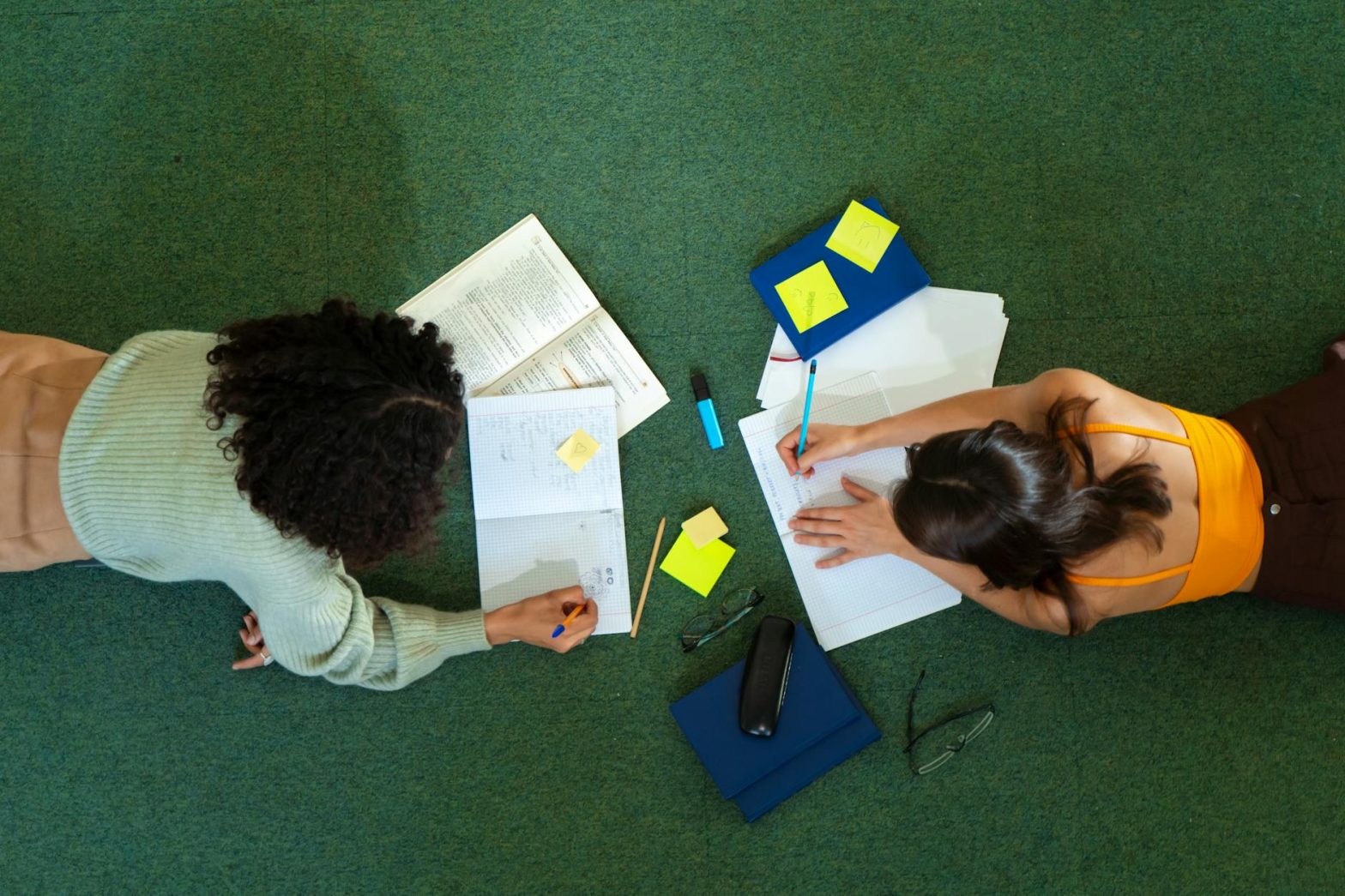 Top view of two women studying on a green carpet, with books and notes, embracing college education.