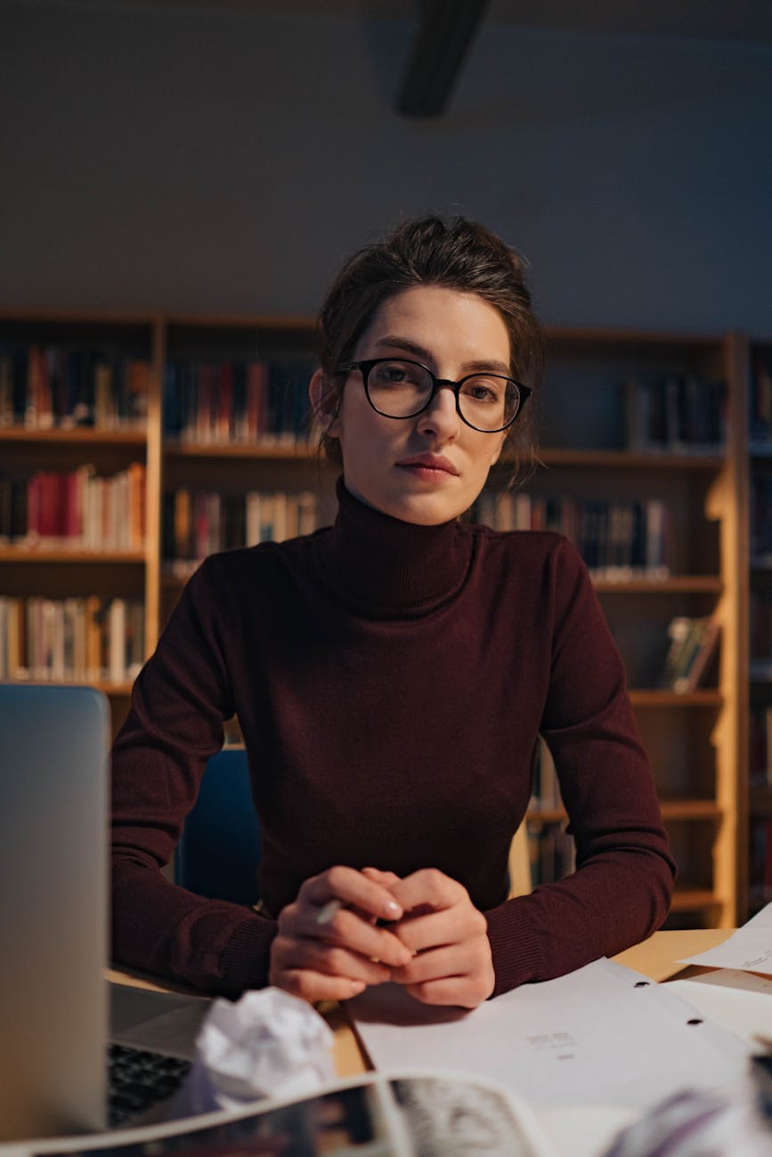 Woman in glasses and long sleeves studying in library, surrounded by books and laptop.