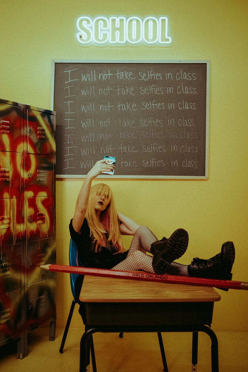 Teenage girl taking a selfie in classroom, defying rules scrawled on blackboard.