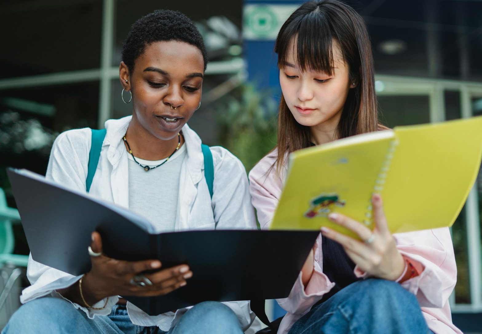 Two young women studying outdoors, focused on their workbooks, showcasing diversity and collaboration.