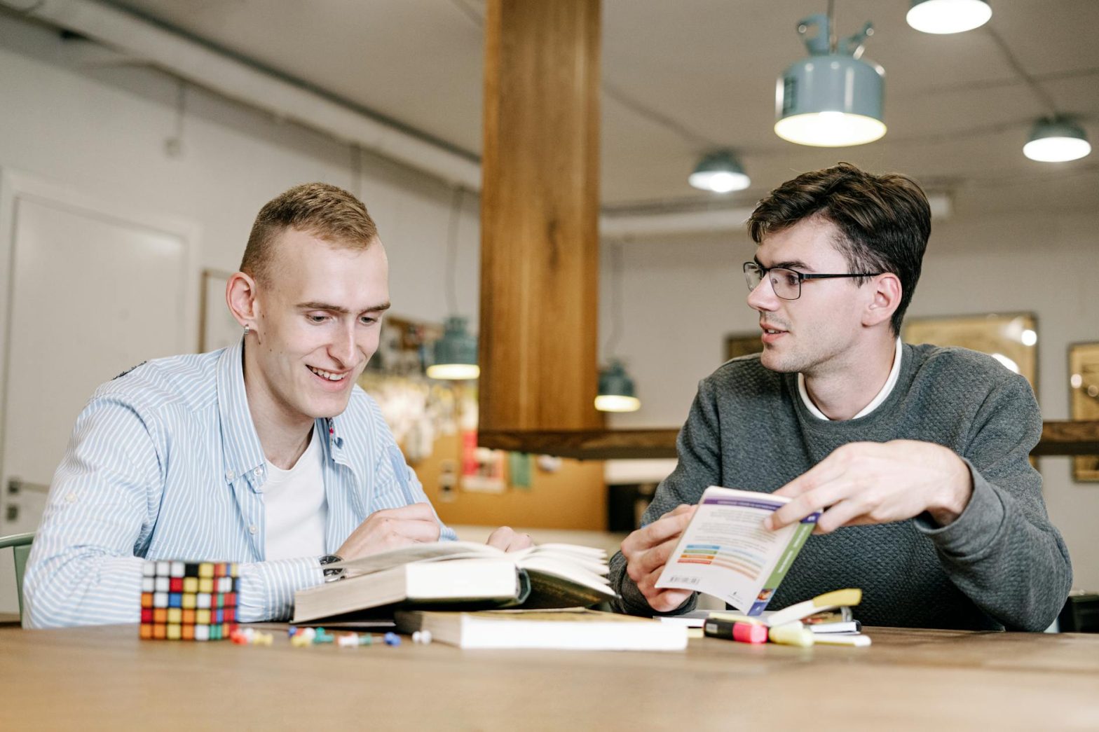 Two young men study together at a table, engaging with books and notes indoors.